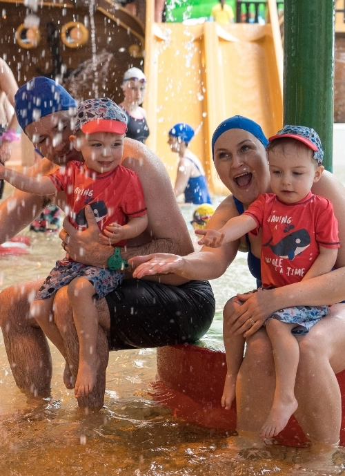 A family sits by a water play area, playfully splashing around. The father and mother, both wearing blue swim caps, each hold a young child in matching red shirts and swim caps. Water droplets surround them as they smile and enjoy the playful moment.