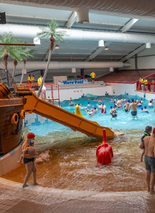 Indoor wave pool with numerous people enjoying the water. A red and yellow waterslide resembling a pirate ship enters the pool from the left. Lifeguards in yellow shirts are stationed around the pool. Fake palm trees and various pool toys are visible.