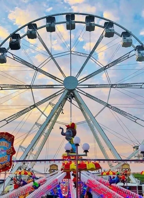 A Ferris wheel and carnival rides are set against a vibrant evening sky filled with clouds. Various colorful balloons and light bulbs adorn the foreground, contributing to the festive atmosphere. A Dumbo-themed ride is visible near the center.