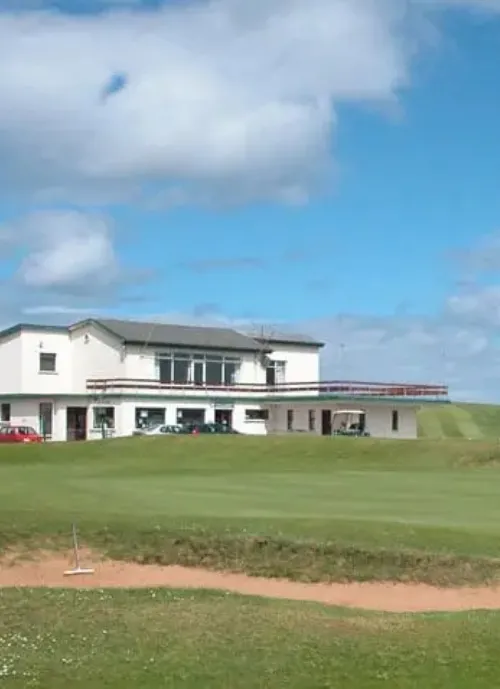 A grassy golf green is in the foreground featuring a sand bunker, with a two-story white clubhouse partially obscured by a small hill in the mid-ground. Several cars are parked beside the building, and the sky above is blue with scattered clouds.