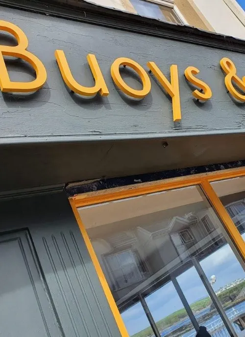 Storefront sign that reads "Buoys &" in bold, yellow letters. The facade below is painted dark gray, and the window reflects a partly cloudy sky and parts of nearby buildings.