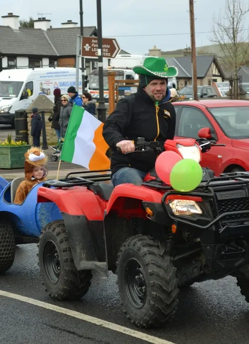 A person in a green cowboy hat rides an ATV adorned with balloons, pulling a small wagon with a child dressed as a brown animal. The ATV has an Irish flag, and the scene appears to be part of a parade with onlookers and buildings in the background.