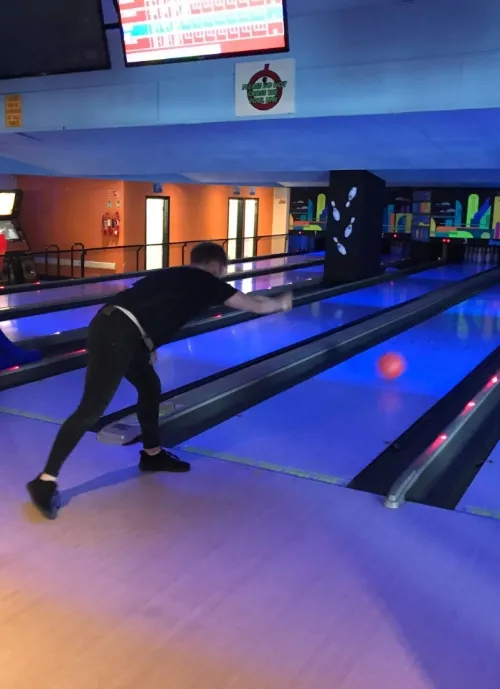 A person wearing a black shirt and dark pants is bowling in a bowling alley. They are mid-action, releasing the ball towards the pins. The alley is lit with blue lights, and the digital scoreboards are visible in the background.