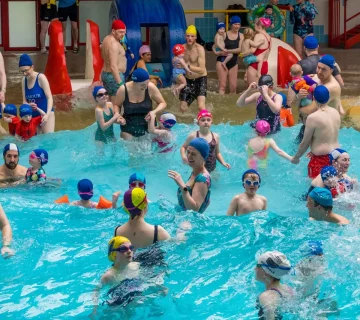 A crowded indoor pool is filled with adults and children wearing colorful swim caps and goggles. Some are playing in the water, while others stand around the edges. Brightly colored inflatables and slides can be seen in the background, creating a lively atmosphere.