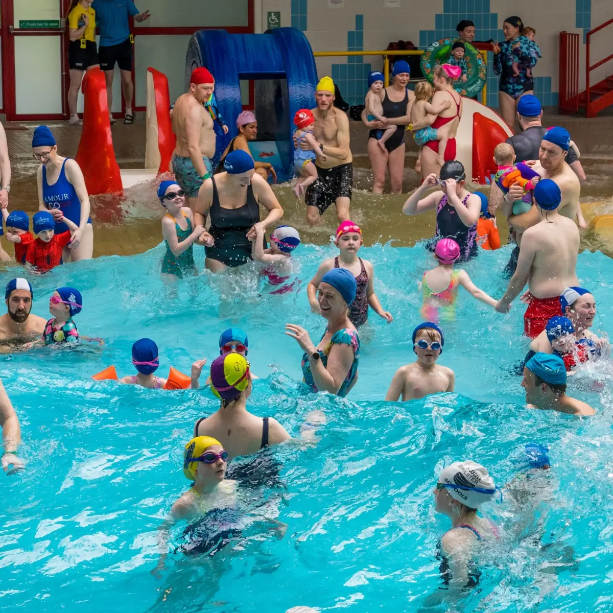 A crowded indoor pool is filled with adults and children wearing colorful swim caps and goggles. Some are playing in the water, while others stand around the edges. Brightly colored inflatables and slides can be seen in the background, creating a lively atmosphere.