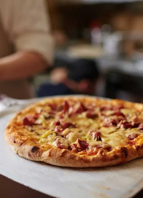 A freshly baked pizza topped with melted cheese and slices of pepperoni sits on a wooden pizza paddle. The background is out of focus, displaying a blurred kitchen setting.