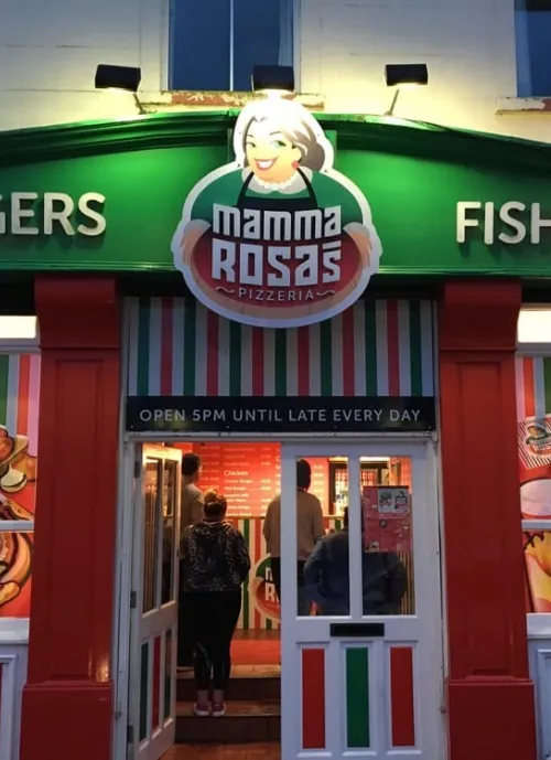 A brightly lit entrance to "Mamma Rosa's Pizzeria" with people standing inside, showing a sign featuring a smiling woman. The facade is decorated with green, white, and red stripes. The door sign indicates that the pizzeria opens at 5 PM daily.