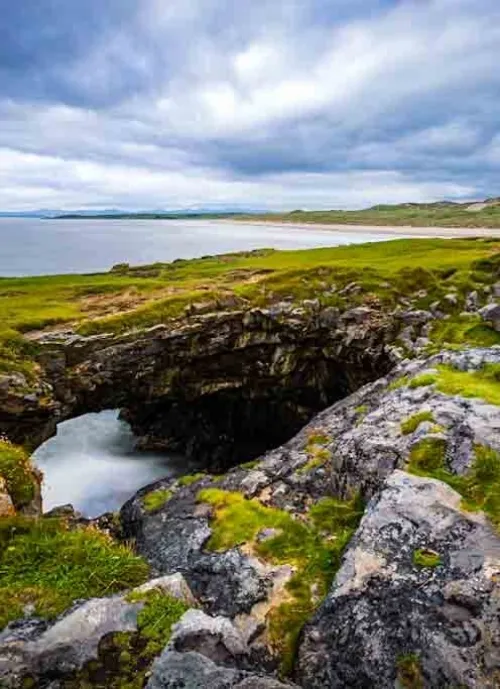 A scenic coastal view shows a natural rock arch formation overlooking the ocean. The landscape features grass-covered cliffs and rugged terrain under a cloudy sky, with waves gently washing against the rocks below. In the distance, a sandy beach meets the sea.