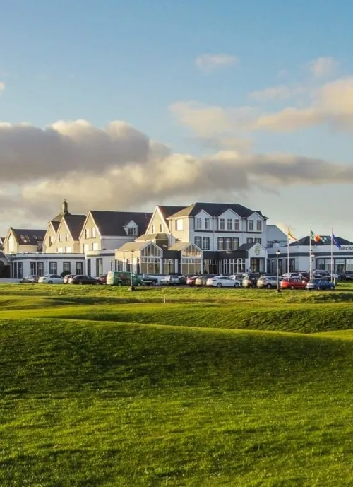 A large, white building with multiple gabled roofs stands against a backdrop of a partly cloudy sky. Several cars are parked in front of it. The foreground features a lush, green grassy area, possibly a golf course or park.