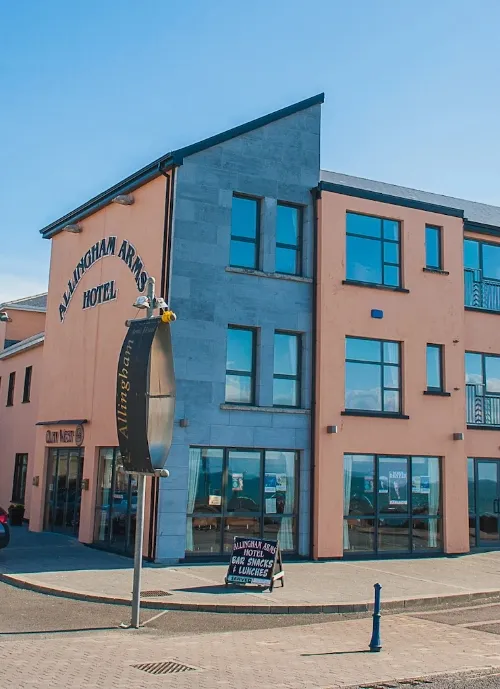 A contemporary three-story building with a sign reading "Allingham Arms Hotel" on its exterior. The facade is partially peach-colored and partially grey stone. There is a round glass entrance and a board outside promoting live music and lunches. The weather is clear and sunny.