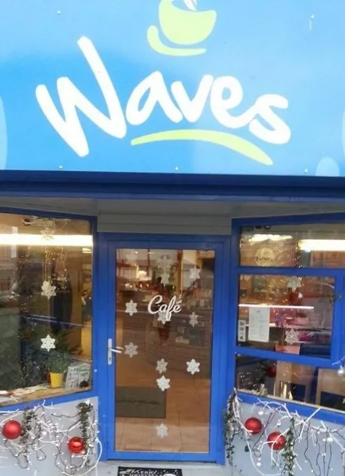 A café with a blue exterior and the name "Waves" written in white and yellow on the sign above the entrance. The glass door has the word "Café" written on it and is decorated with white snowflakes. The entrance is adorned with Christmas decorations including baubles and lights.