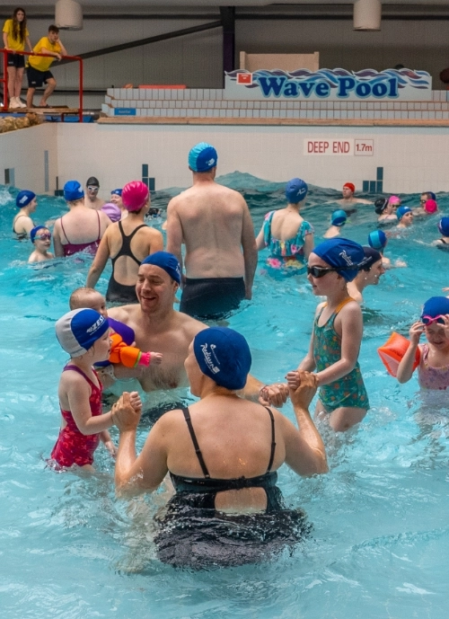 A crowded indoor wave pool with adults and children wearing swimsuits and swim caps. Some people are holding hands or playing in the water. A sign reads "Wave Pool" and "Deep End 1.7m" above the pool. The atmosphere is lively and engaging.