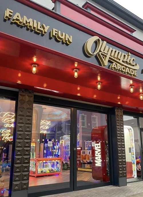 Storefront of the "Olympic Arcade" with a large red marquee and golden letters reading "Family Fun" and "Olympic Arcade." The entrance displays bright neon lights and signage inside, with arcade games visible through the glass doors.ibrant daytime setting.