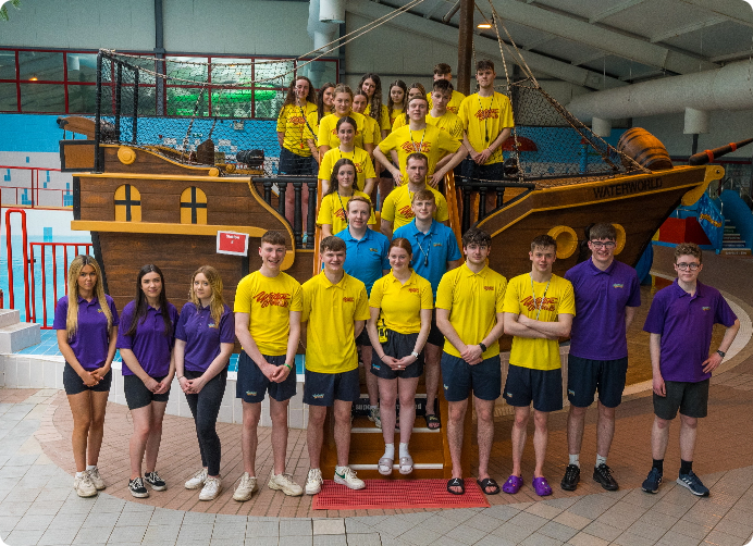 A group of young people stand in front of a wooden ship structure inside a building. The group is divided into two sections: those in yellow shirts stand on the ship, while those in purple shirts stand on the ground. Some people are smiling, and others are neutral.