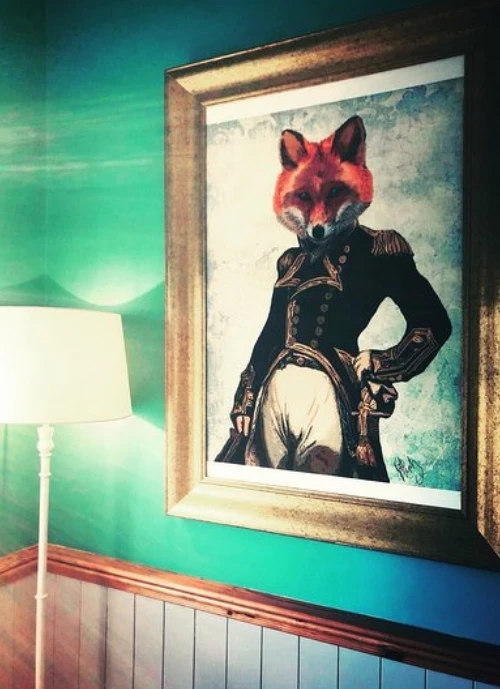 A framed artwork of a fox dressed in a classic military uniform with epaulettes on its shoulders hangs on a teal and turquoise wall. A white floor lamp is positioned to the left of the picture.