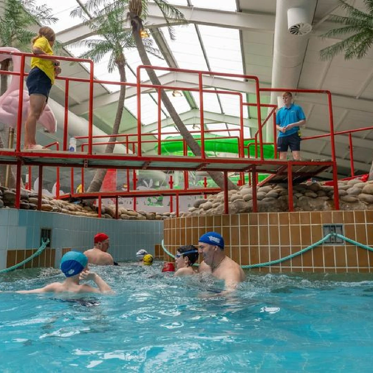 People are swimming in an indoor pool with red railings and palm trees in the background. Two individuals, one in a yellow shirt and one in a blue shirt, are supervising from above. Swimmers, including children and adults, are wearing swim caps.