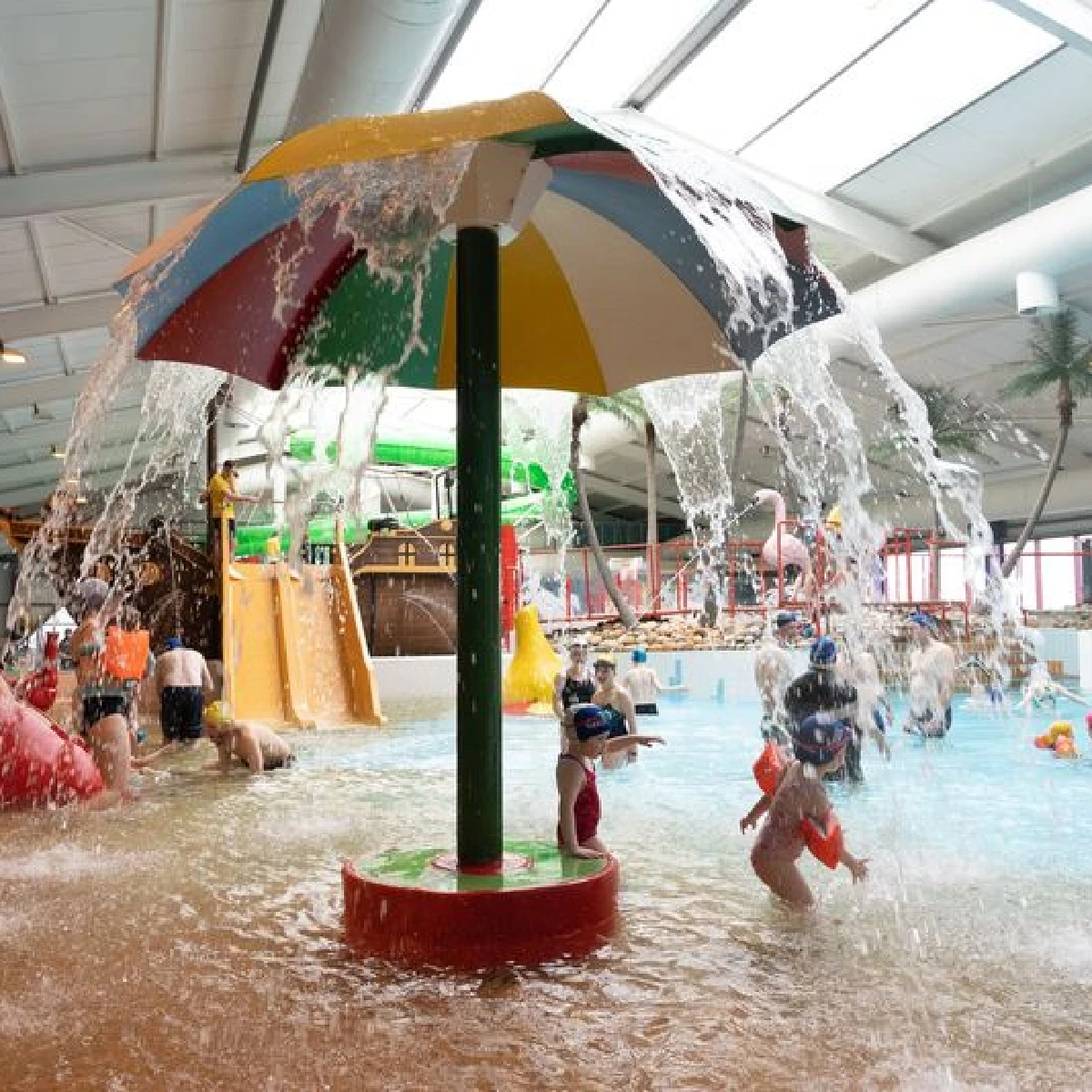 Children play under a colorful water fountain shaped like an umbrella at an indoor water park. The scene has slides and various aquatic structures, with kids enjoying the water activities. The atmosphere is lively and playful.