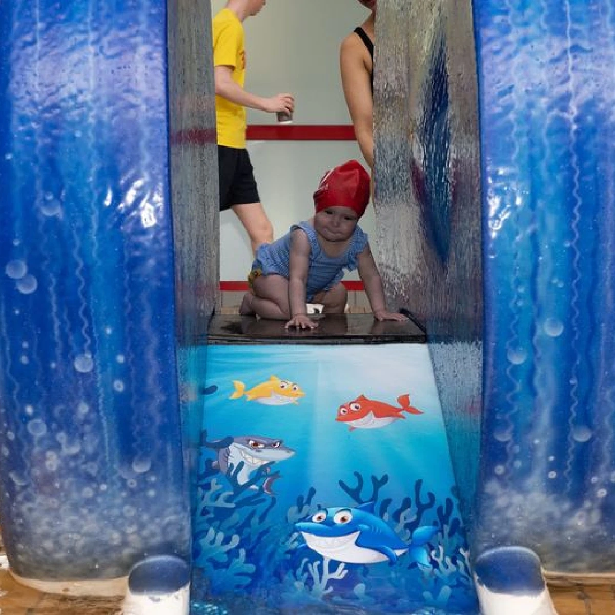 A child wearing a red swimming cap and blue swimsuit crawls inside a blue water slide tunnel decorated with colorful fish graphics. An adult and another child in the background stand near the entrance. The scene is part of a water park.